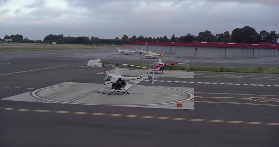 Private planes and helicopters are parked on the tarmac of a small airport at dusk on a cloudy day