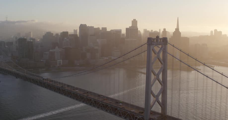 Boat wake passes under suspension bridge near downtown as fog shrouds skyscrapers and sun rises on horizon