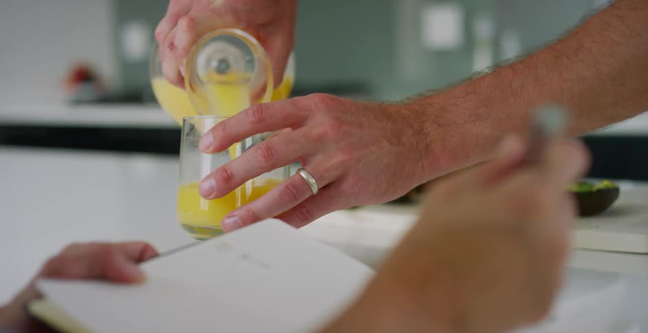 Husband picks up glass off of kitchen counter to pour orange juice as his wife goes over her notes in a notebook