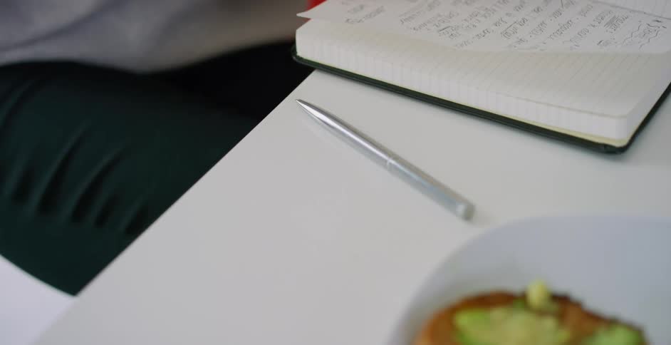 A white person sits at a table with a notebook and a half eaten piece of toast on a plate