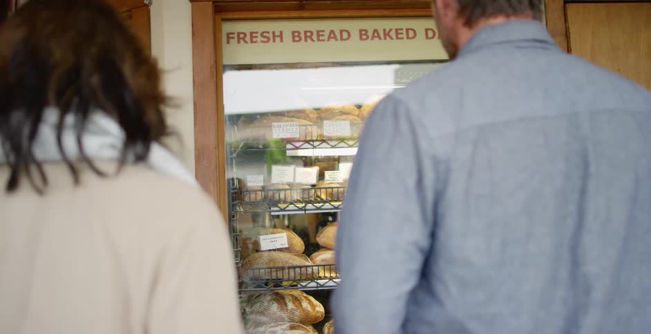 Man with a blue shirt and woman wearing a white scarf and trench coat stand and look at the bakery window display