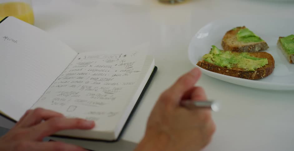A woman sits at the kitchen counter and looks at her notebook while a man makes them avocado toast