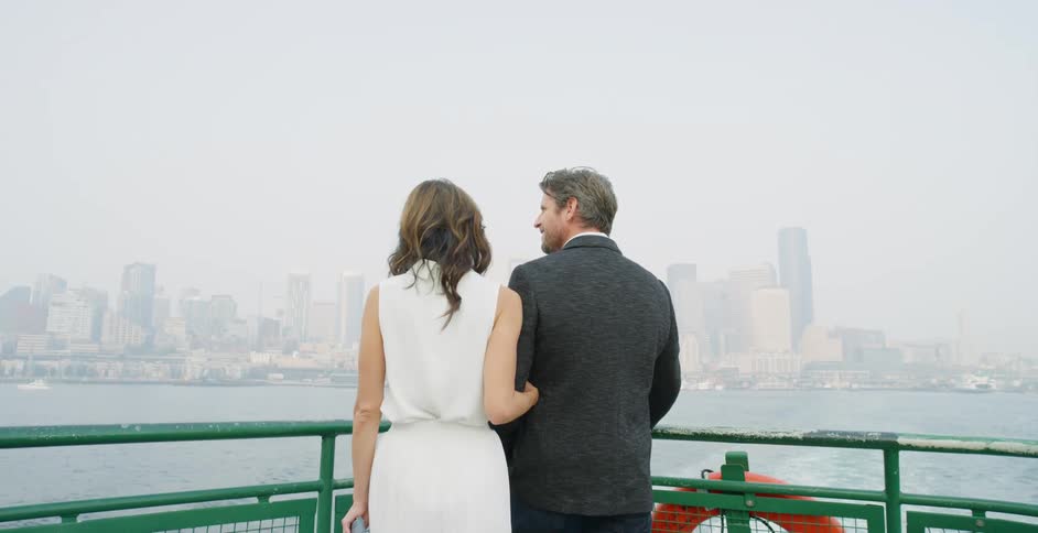 A man and woman arm in arm stand on a boat and look towards the city skyline