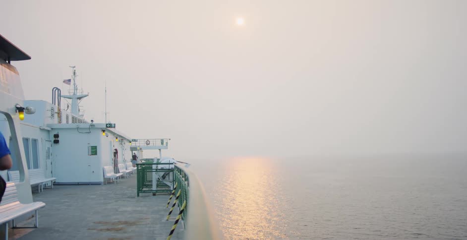 A man is walking on the deck of a large ship as it travels on the ocean on a foggy day at dusk