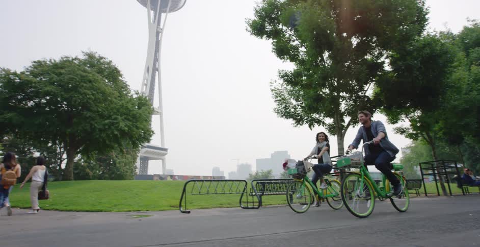 Man and woman ride bicycles past the park benches as pedestrians walk nearby