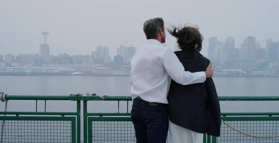 A man puts his arm around his wifes shoulders as they stand on a pier and take in views of the ocean and city skyline