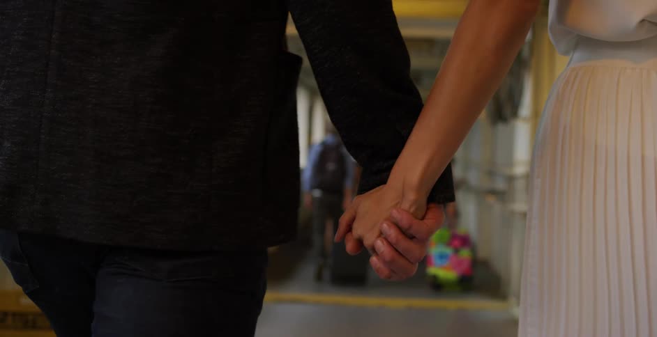 A man and woman holding hands walk down a corridor behind a family pulling suitcases