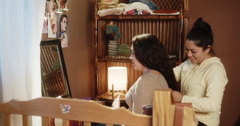 A woman laughs as she brushes her sisters hair as they sit in front of a mirror in a bedroom