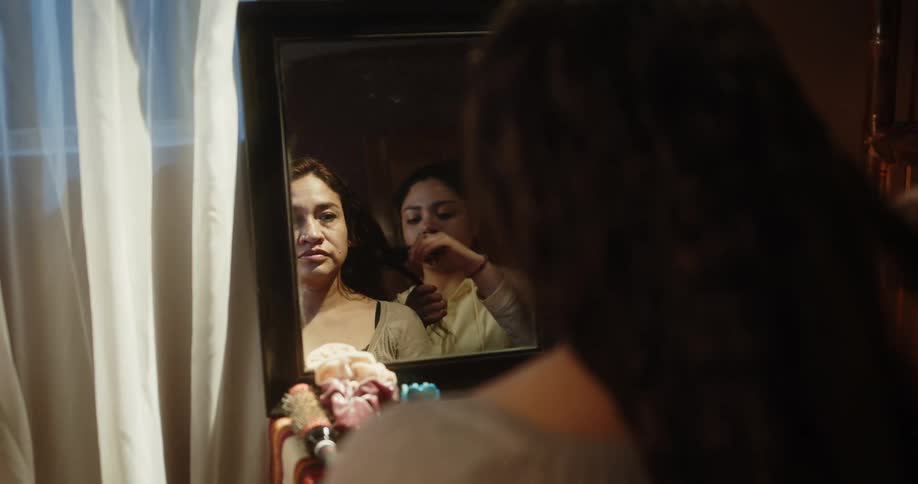 A woman combining another womans hair in front of a mirror next to a window