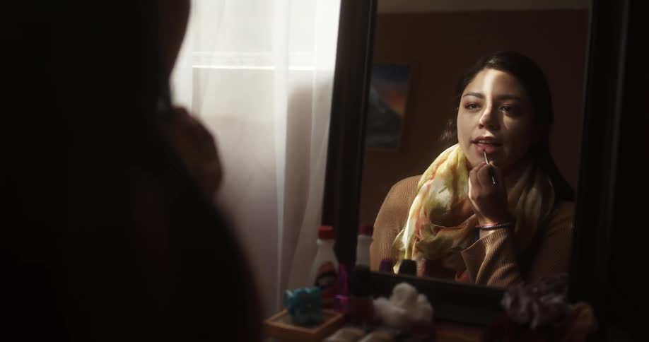 Brunette woman with a scarf sitting on a stool and looking in a mirror puts on lipstick