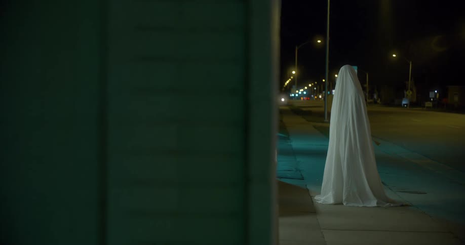 A figure covered in a sheet stands on a city sidewalk in front of a building
