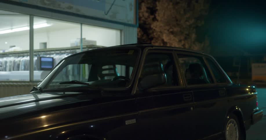 An empty black car parked outside a building with its headlights on at night