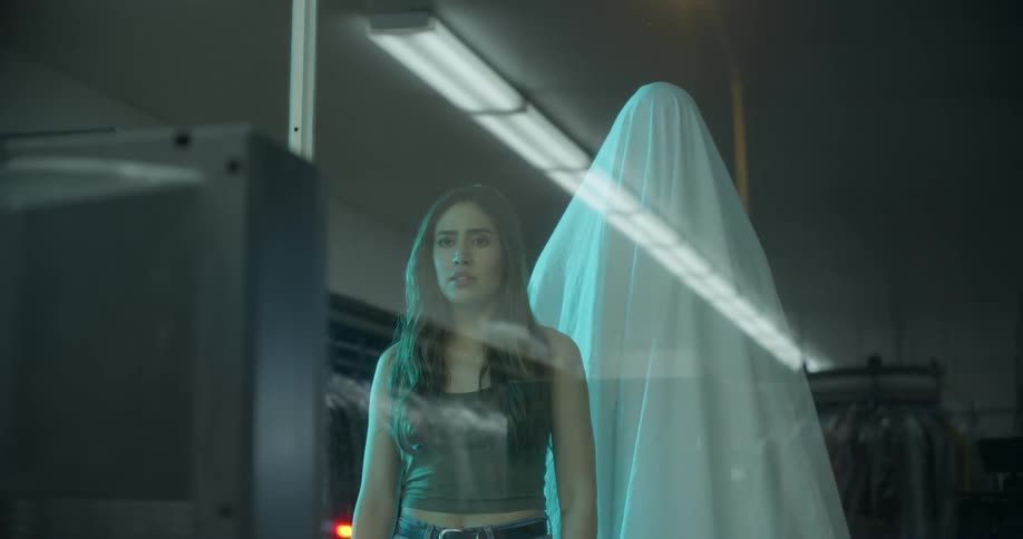 Ghost stands behind a woman watching television through the display window of a store at night