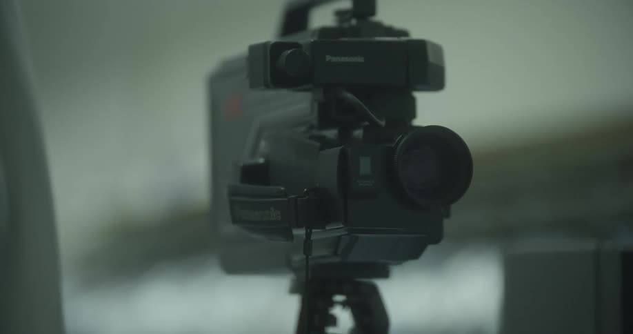 An old style video camera sits on a tripod next to a television showing static