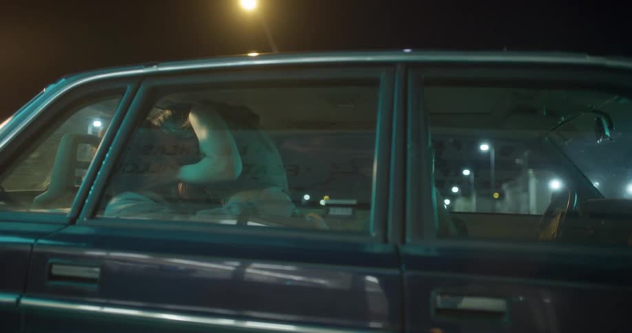 Man and woman kiss passionately on the backseat of a car parked on a parking lot at night