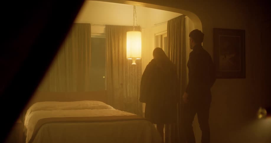 A woman begins to remove her coat as she approaches a bed with a man in a dimly lit room