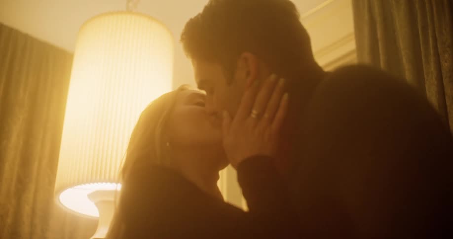 A man and woman kissing indoors while standing next to a pair of windows