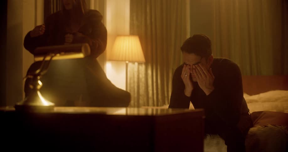 A man sitting on the end of a bed wipes his eyes as a a woman puts on her coat and walks away