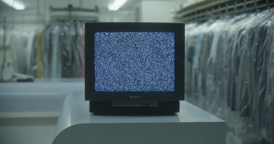 A television set on a counter of a dry cleaning store with clothing hanged up in the background