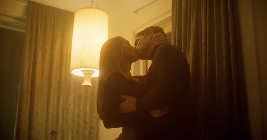 A latin man and woman kiss and embrace beneath a hanging light in a room