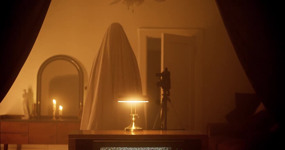Figure covered in a bedsheet stands behind a vintage television showing static in a room