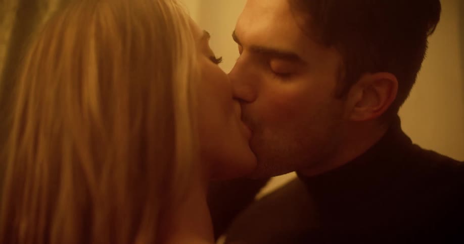 Man with a black turtleneck sweater and blonde woman kiss each other passionately in the bedroom