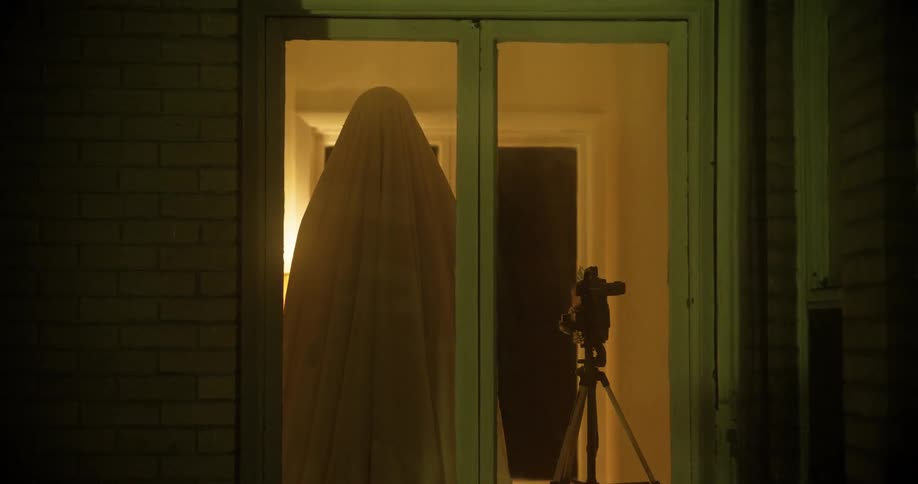 Figure draped in blanket stands inside glass door next to video camera