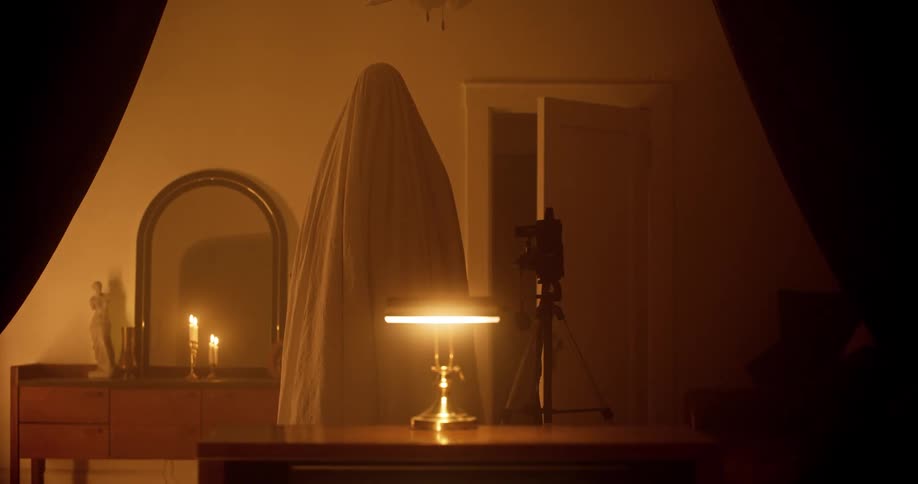 A person stands under a sheet like a ghost in a dim bedroom
