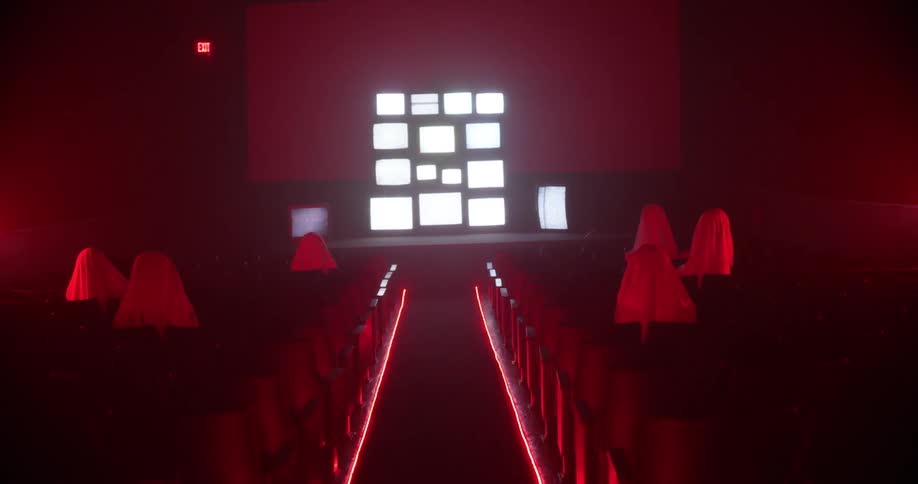 People covered in white sheets sit in a room with a bank of monitors on the wall flashing static