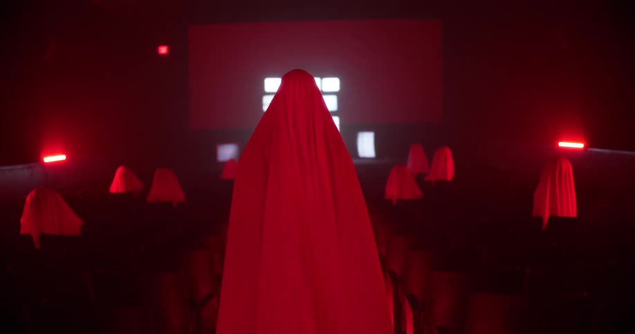 Person wearing a sheet walks down a theatre aisle while other sheet covered people sit in the seats