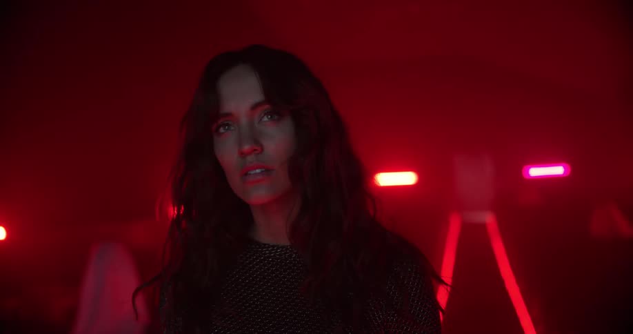 Woman staggers to stop in aisle of red tinted theater occupied by shrouded figures