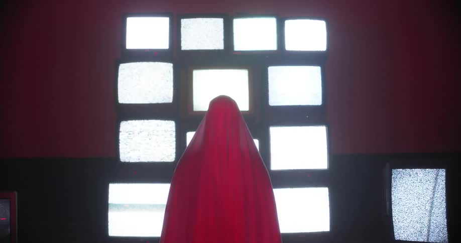 A person with a red cloak stands in front of a wall of televisions with fuzzy screens