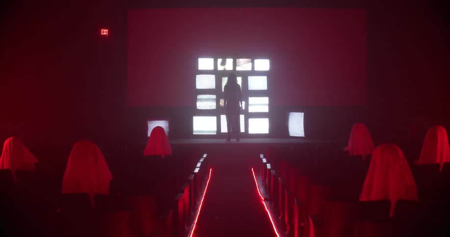 A person stands at the front of a room in front of a wall of televisions