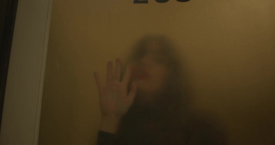 A white woman dances seductively with her face and hands pressed against glass in a door