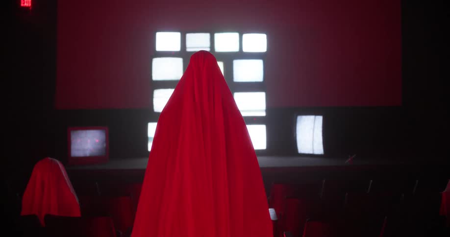 A figure dressed entirely in red walks to a stage covered in television sets playing static