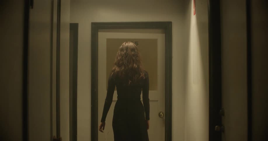 A young woman walks and stops in front of a door that has a red light flashing behind it