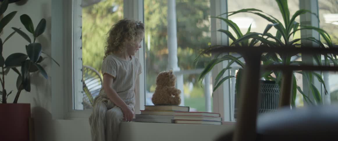 A little girl with a teddy bear walking up and sitting by a window