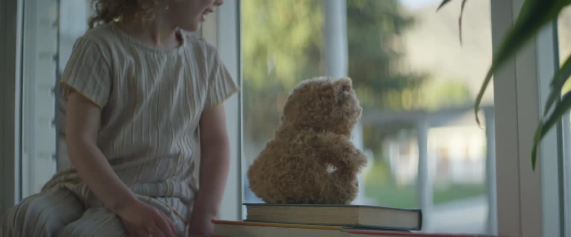 Young girl places teddy bear on stacked books facing out toward window