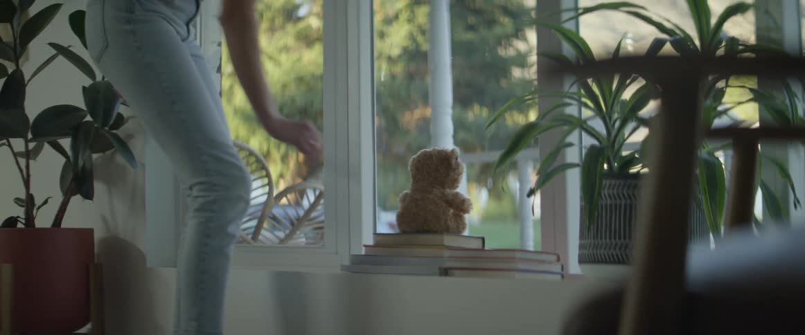 A teenager puts a teddy bear on some books by a window in her house