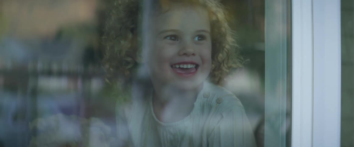 A young girl with curly hair smiles as she looks out the window of her house