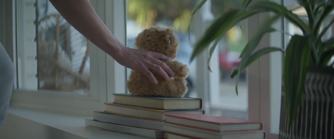 A white woman sets a teddy bear on a stack of books on a shelf by a window