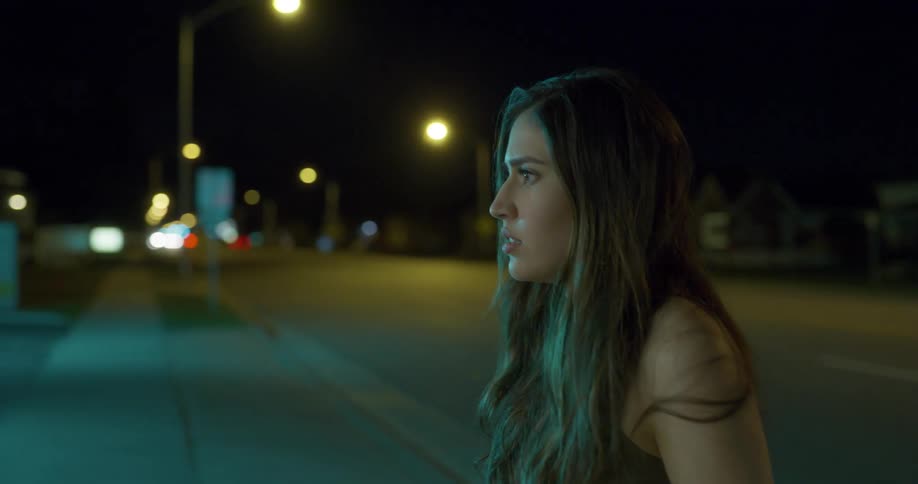 Woman steps out of a car parked on the street of a small town at night to look through a shop window