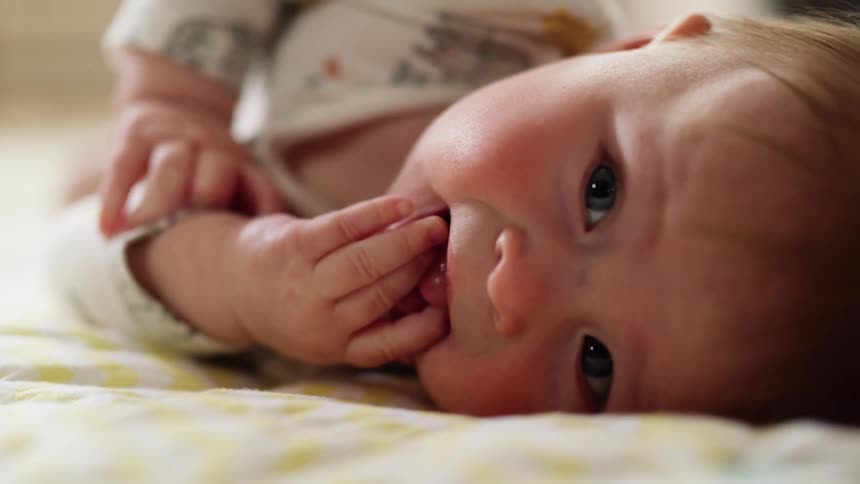 Baby laying on a bed puts his fingers on his mouth and smiles
