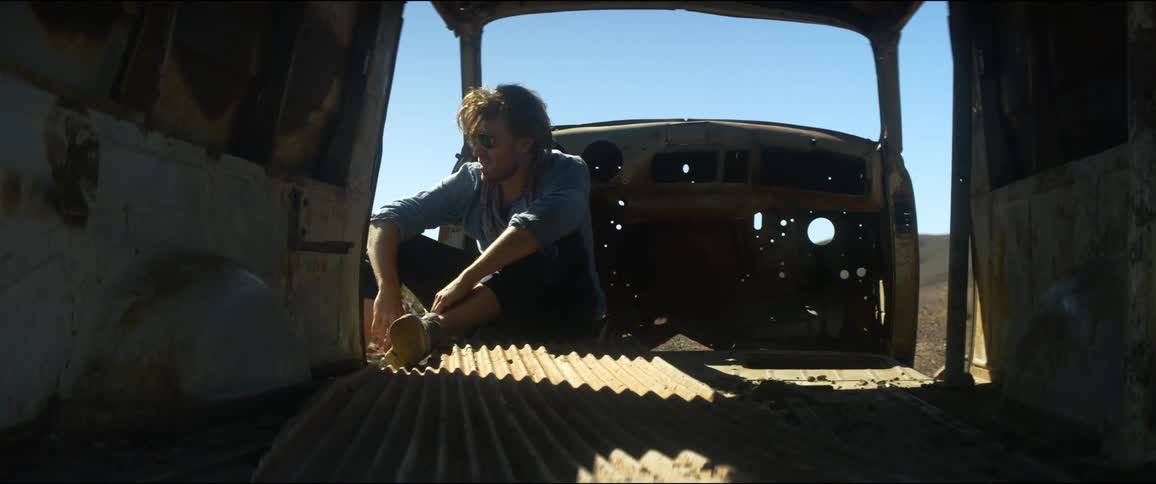 A white man sits inside of a wrecked vehicle out in the desert