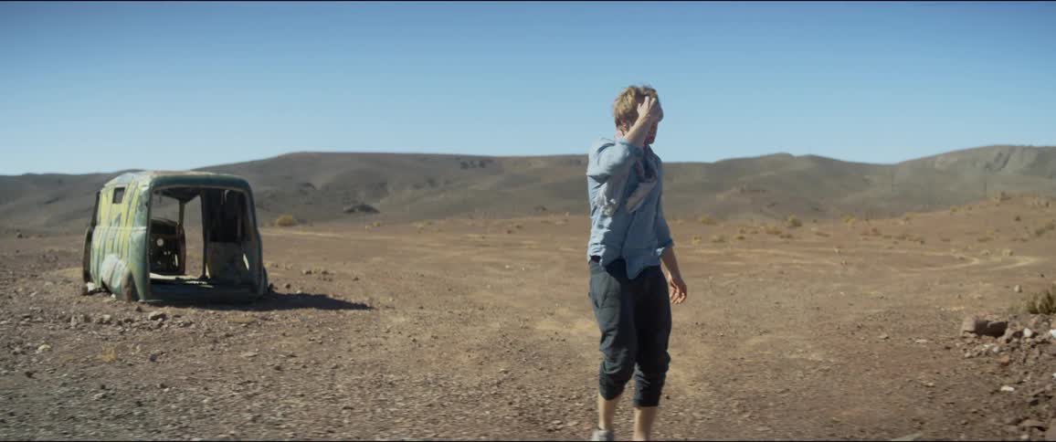 A man in sunglasses walks through a barren landscape towards the dilapidated frame of a minivan