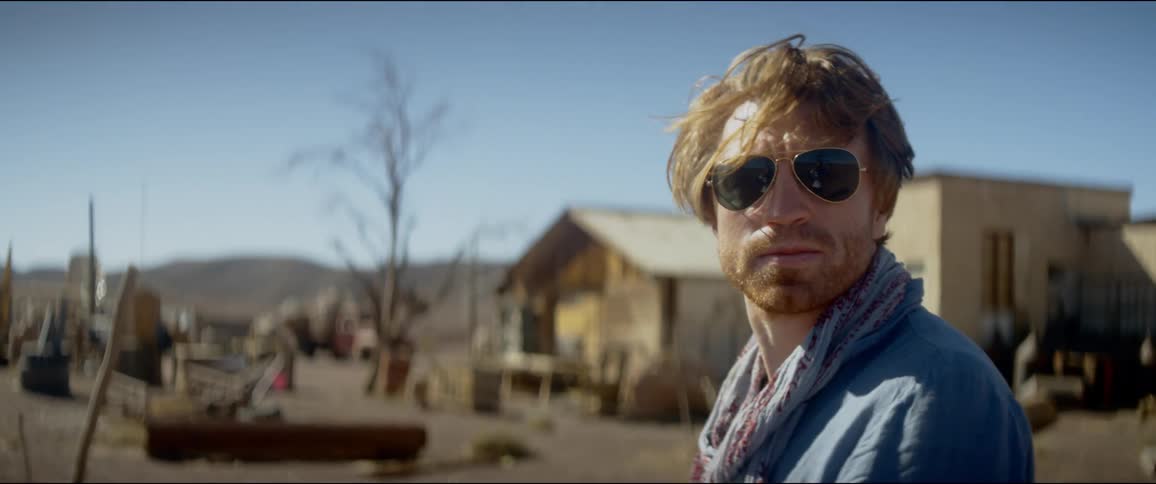 Blonde man wearing sunglasses walks around a rural area