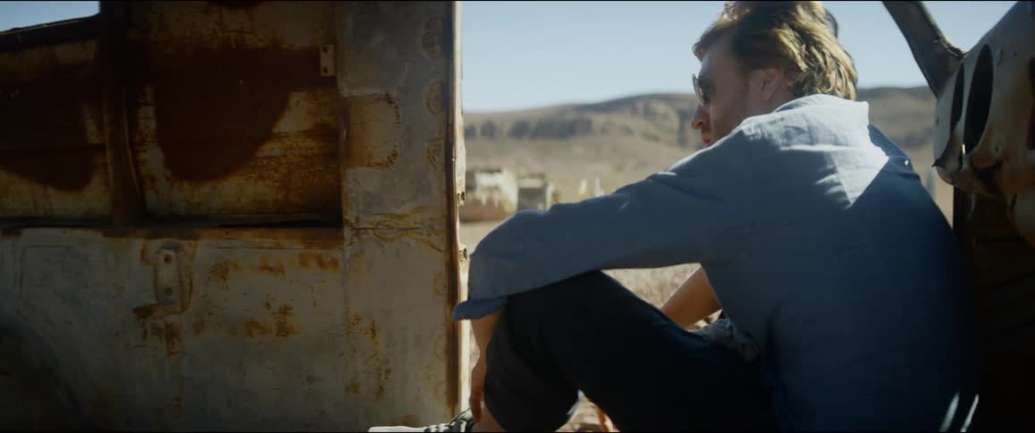 A man sits in the rusted shell of a stripped truck as the wind blows his hair and shirt