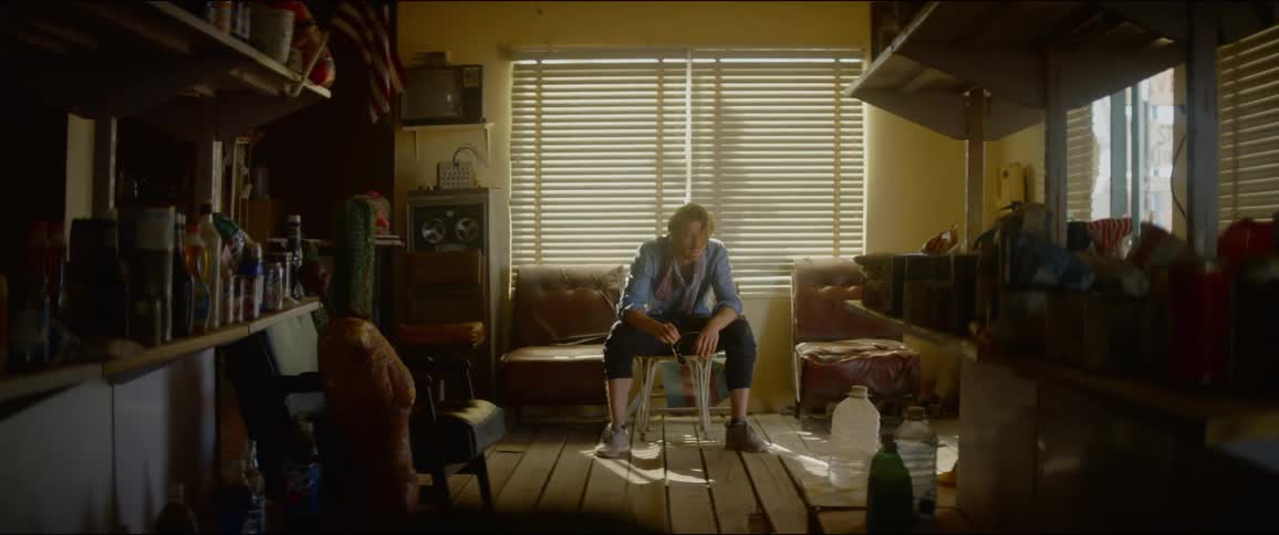 A disheveled man sits on a chair in his cluttered and messy home