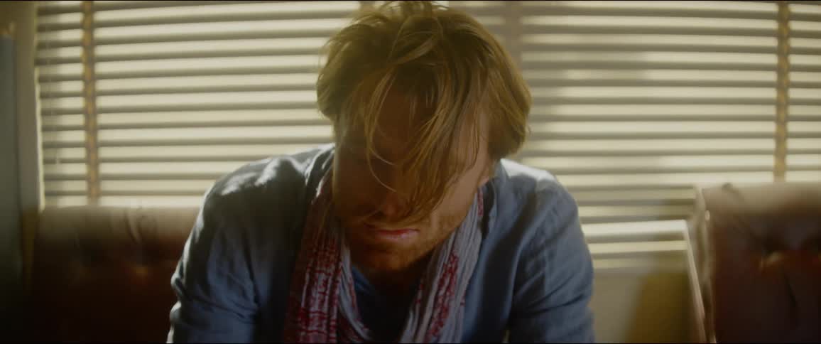 A man sits on his couch with his head down as wind blows through his hair