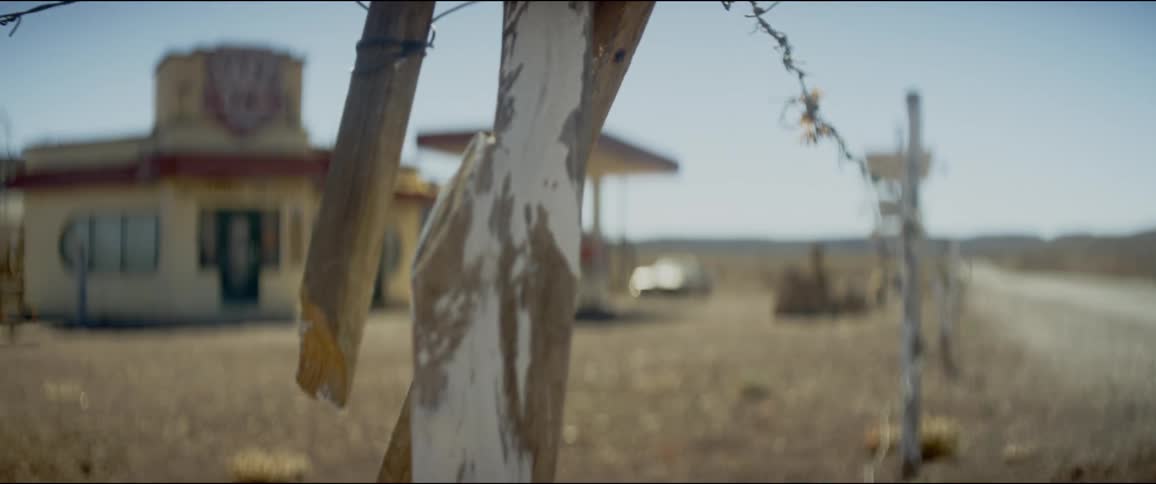 Broken wooden posts hang from the barbed wire of a fence between the road and a rural gas station and store
